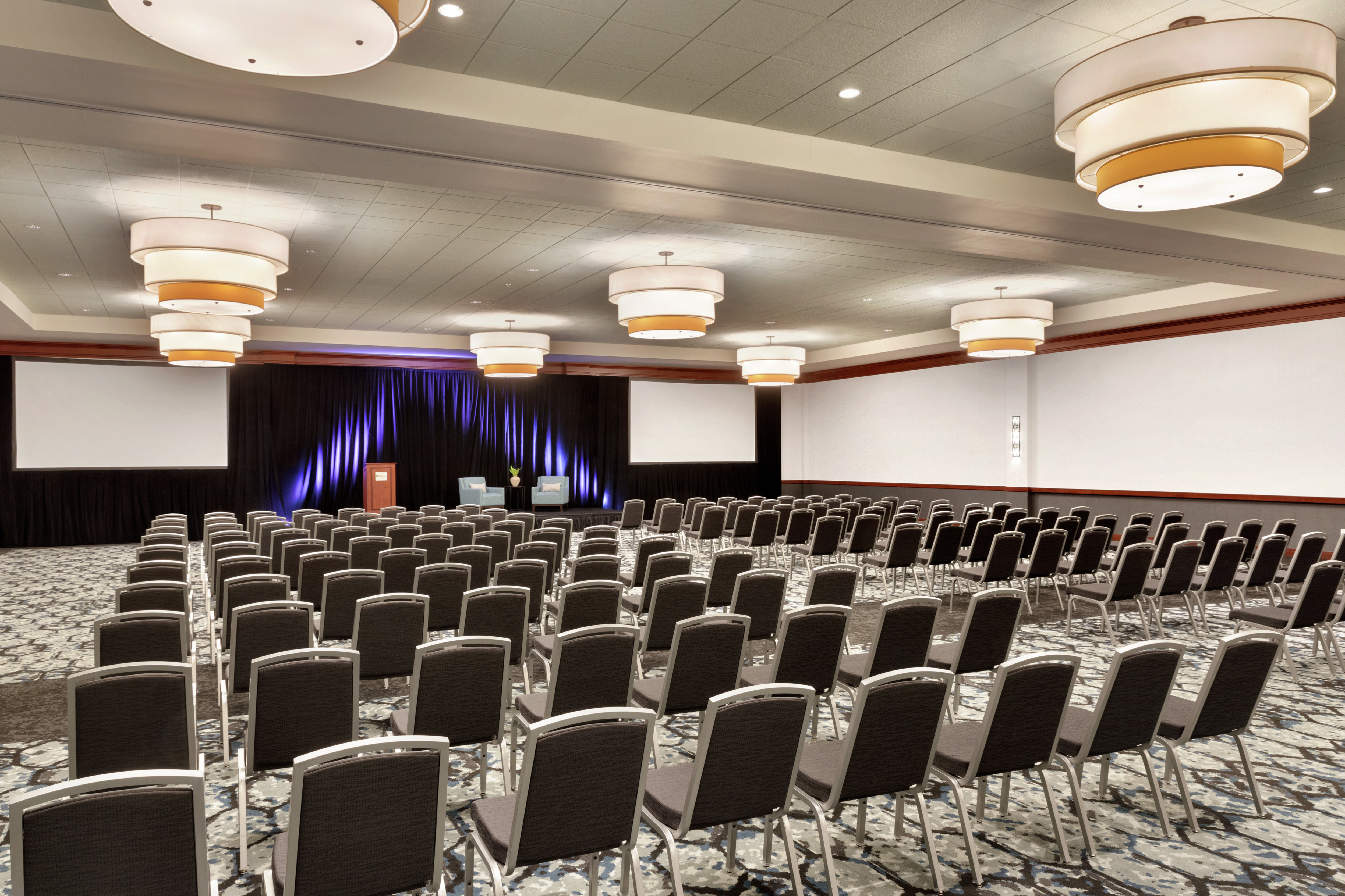 Spacious hotel ballroom featuring theater setup and stage with podium at front of room.