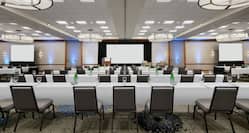 Spacious ballroom facility equipped with classroom style tables, projector screens, podium, and stage.