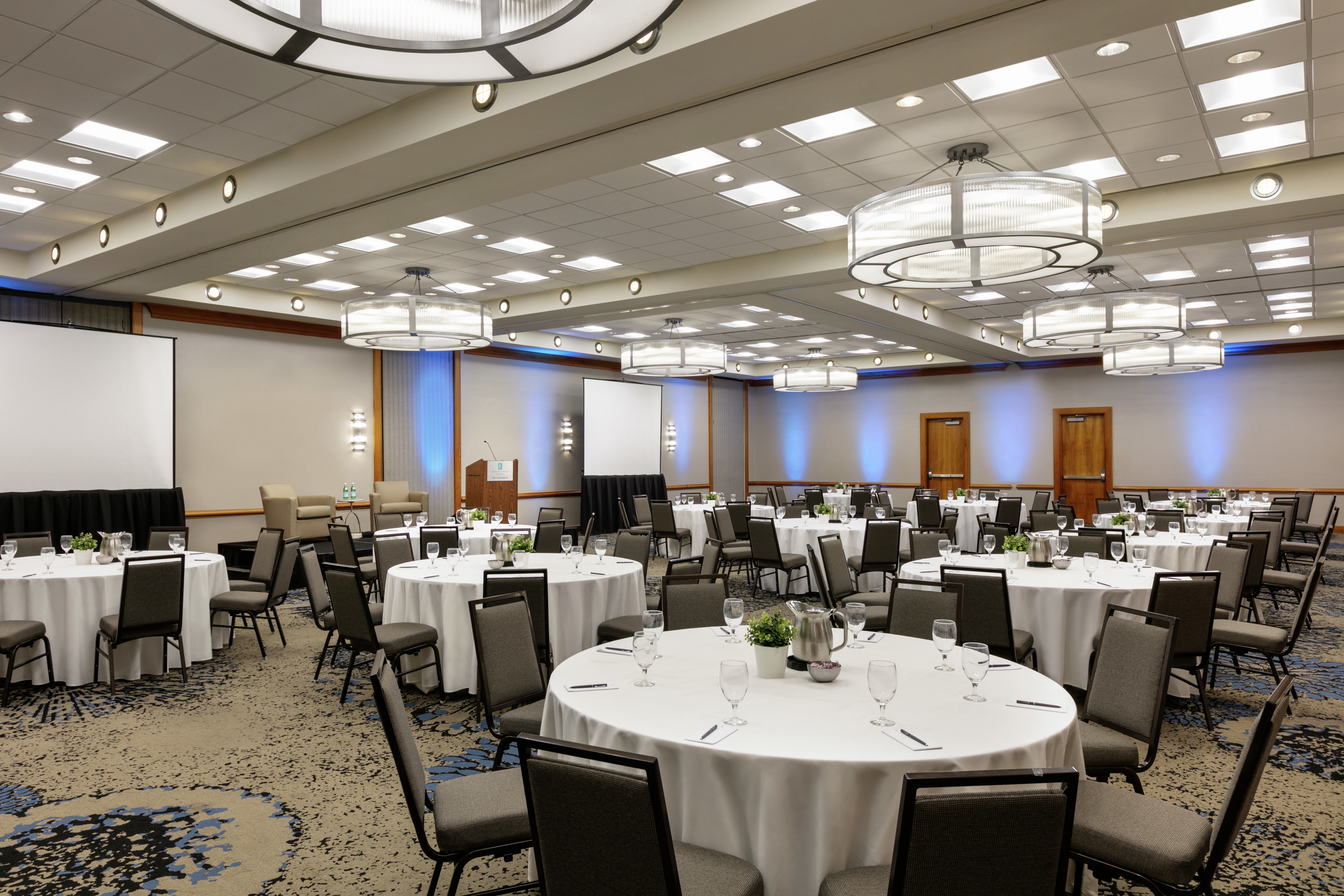 Spacious ballroom facility equipped with round tables with beautiful place settings, project screens, and stage.