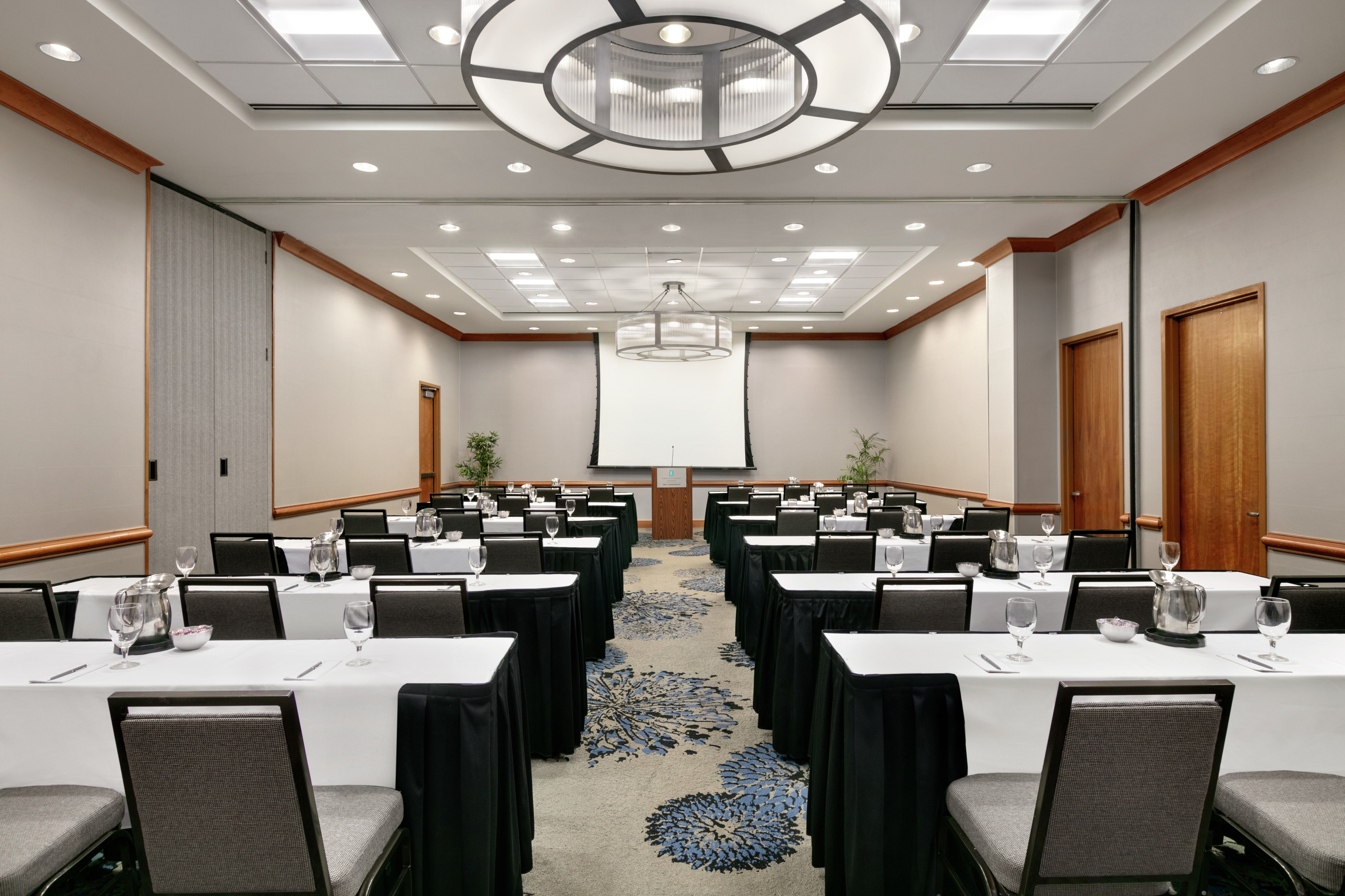 Spacious meeting room facility equipped with classroom style tables, projector screen with equipment, and podium.