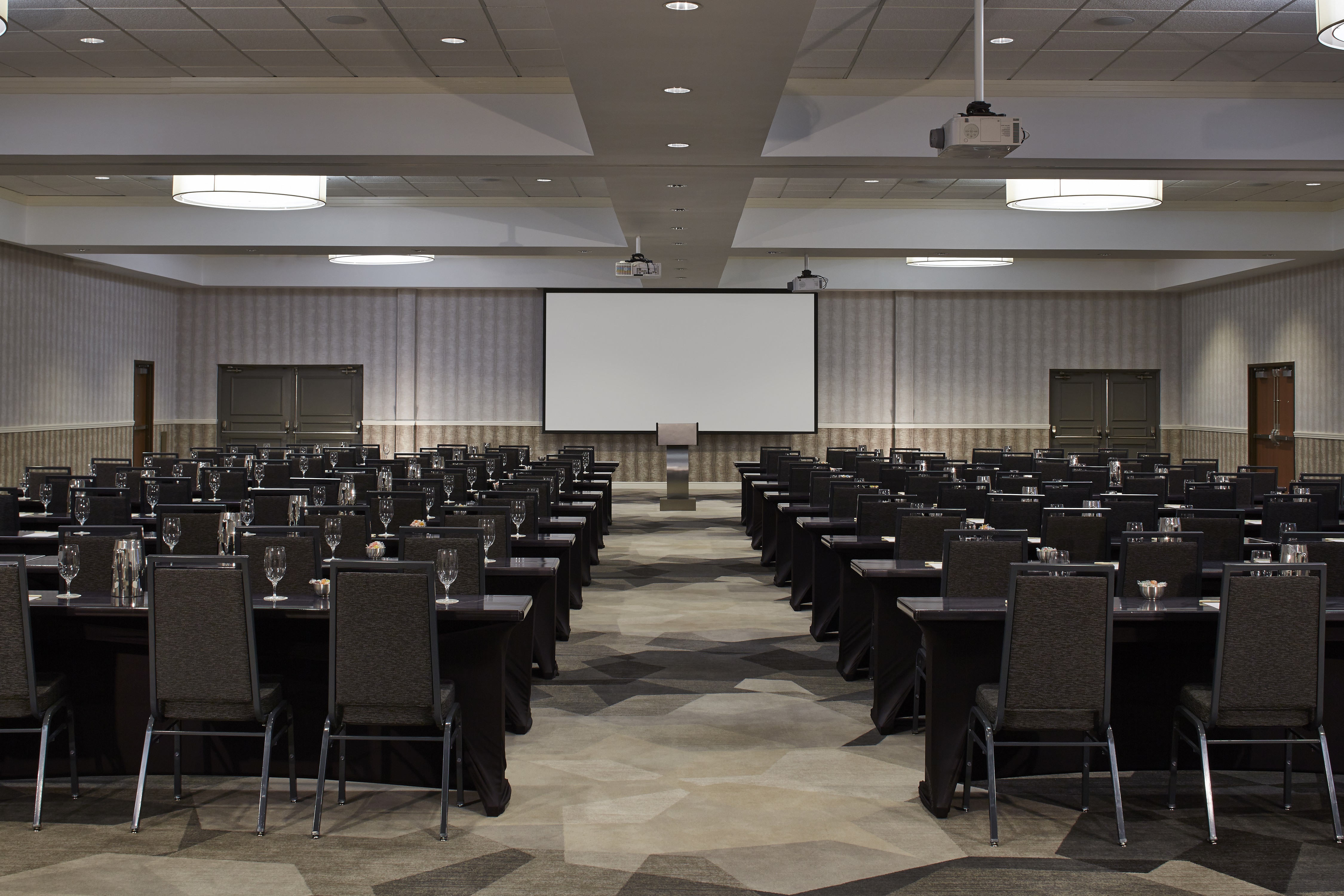 Classroom Setup in Meeting Room With Projector Above Tables and Black Chairs Facing Podium and Presentation Screen