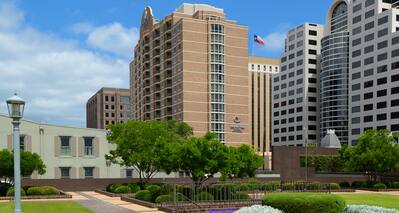 Daytime View of Hotel Exterior, Signage, Flagpole, and Landscaping From Grounds of Texas State Capitol 