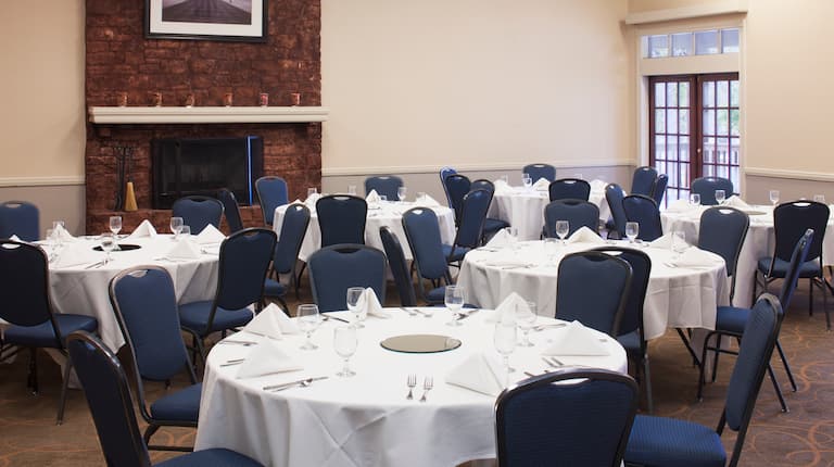 Place Settings on Round Tables With White Tablecloths, Blue Chairs, Wall Art Above Fireplace, and Window in University Meeting Room 