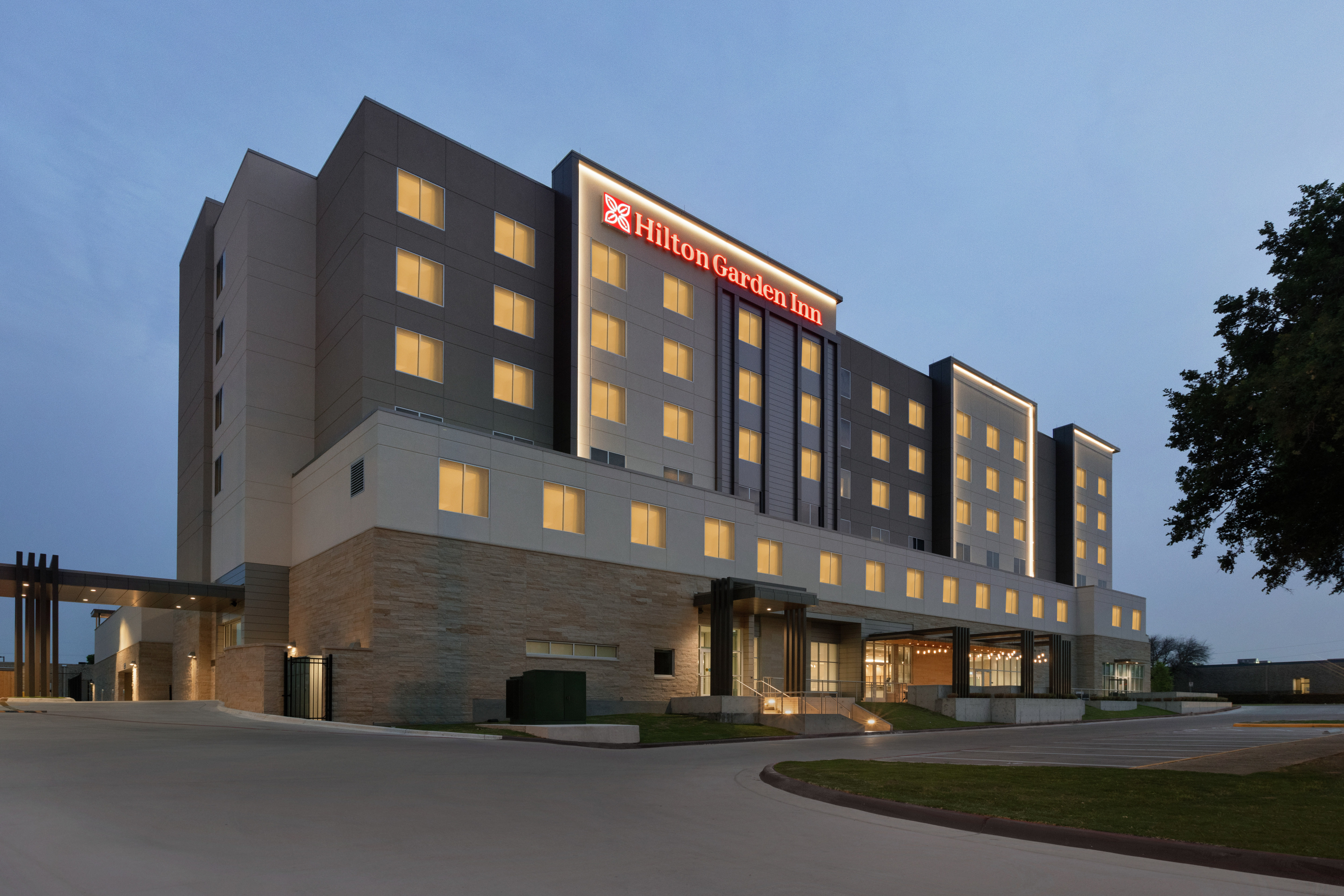 Front view of modern Hilton Garden Inn hotel featuring flowing guest room lights and clear dusk sky.