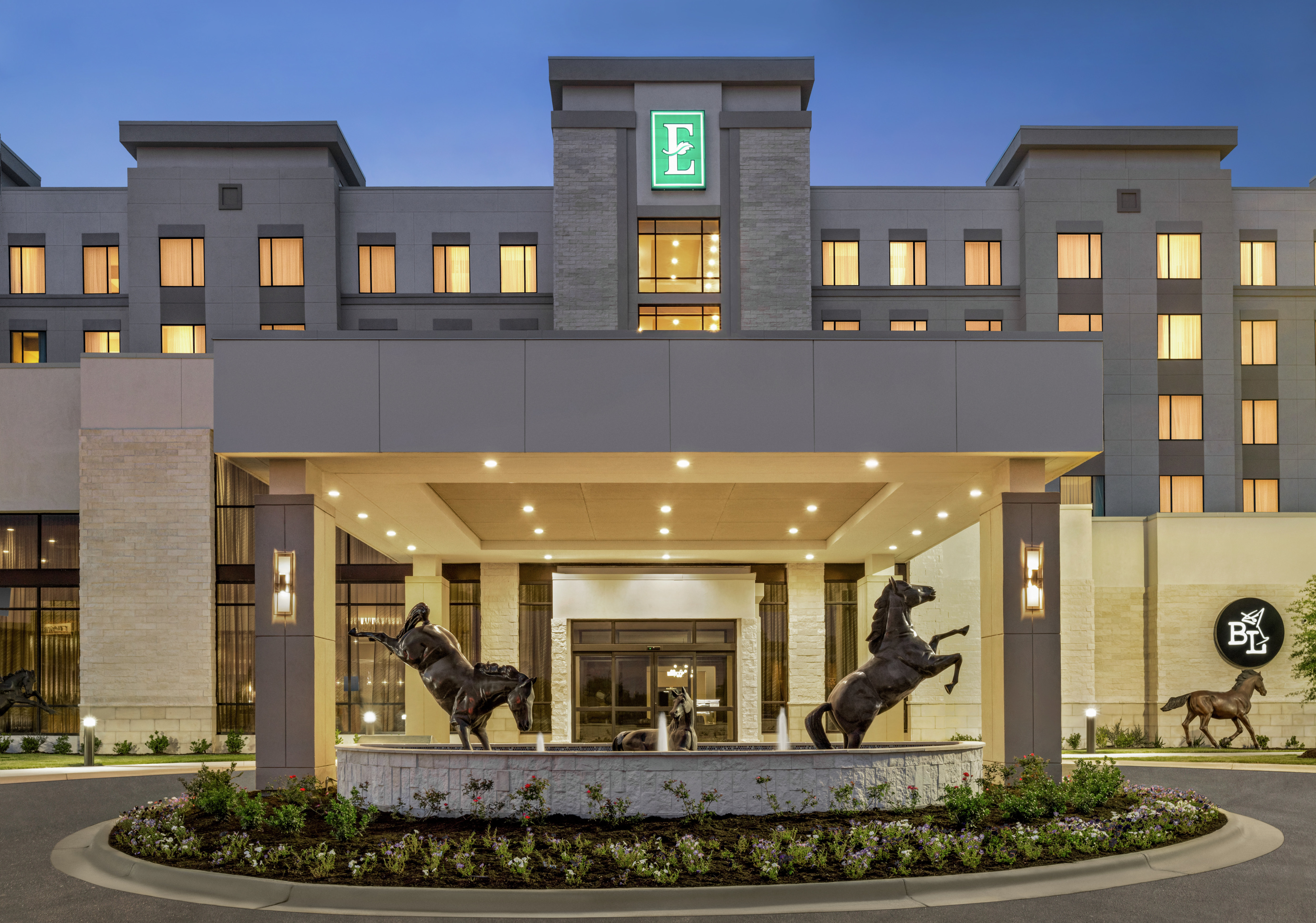 Modern Embassy Suites hotel exterior featuring porte cochere, stunning horse statues, and glowing guestroom windows.