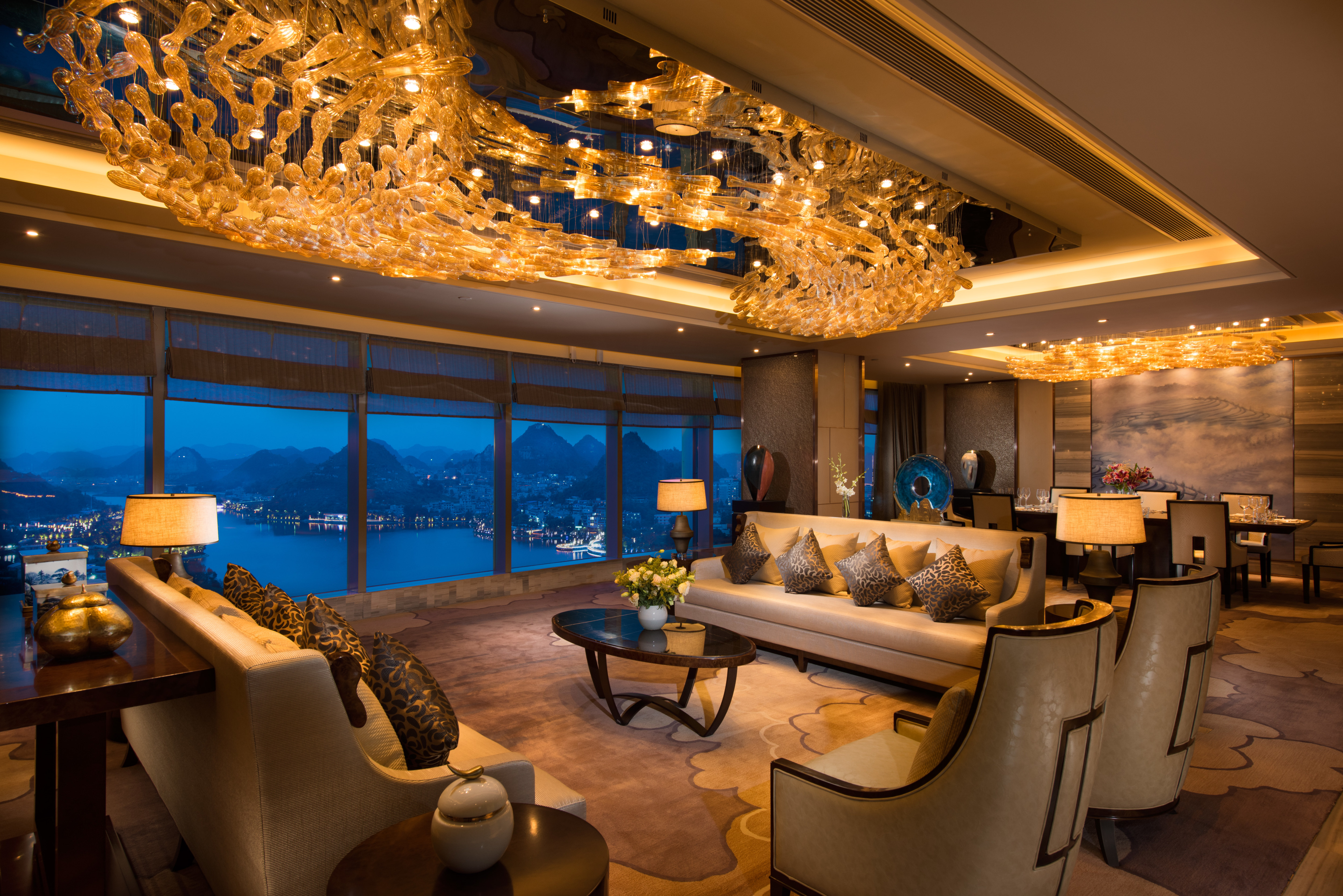 Illuminated Presidential Suite With Tables, Soft Seating, and Large Window With Lake View at Night