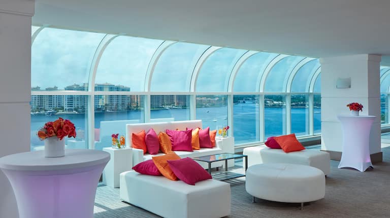 View of Couch, Ottoman and Additional Seating with Magenta and Orange Pillows, and Tables with Floral Arrangements in the Atlantic Ballroom Lounge that Overlooks Water
