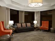 Sofa Between Two Illuminated Lamps on Side Tables, Two Orange Armchairs, and Ottomans by Windows With Open Drapes
