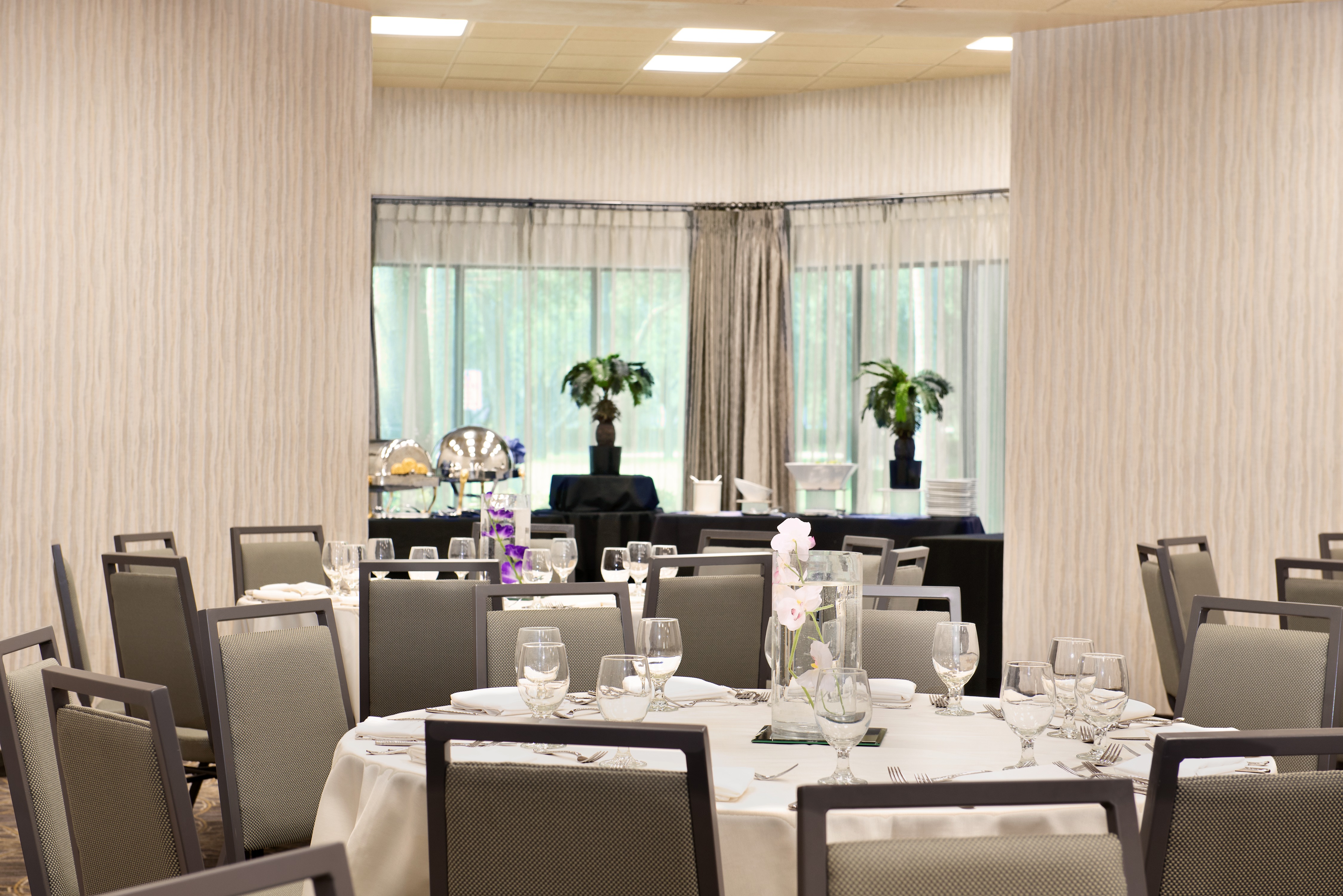 Place Settings, Drinking Glasses, and Flowers on Dining Table With White Linens, and Food Service Area by Window With Long Drapes in Bermuda Room