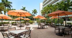 Daytime View of Tables With Orange Umbrellas and Chairs, Outdoor Pool, and Hotel Exterior Surrounded by Palm Trees