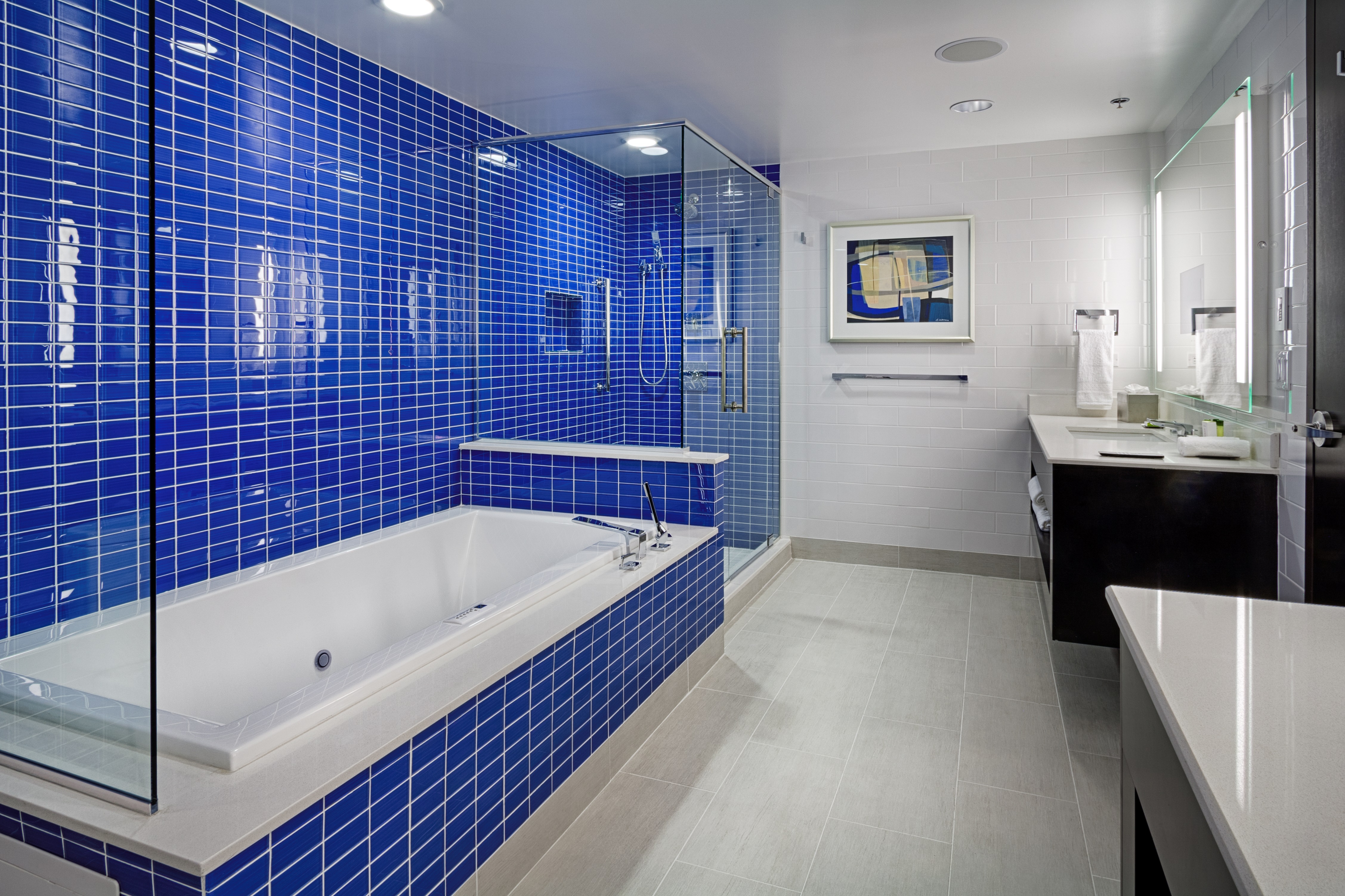 Bathroom With Blue Tile Surrounding Large Tub and Shower With Glass Door, Wall Art, Brightly Lit Vanity Mirror, Sink, Fresh Towels, and Amenities