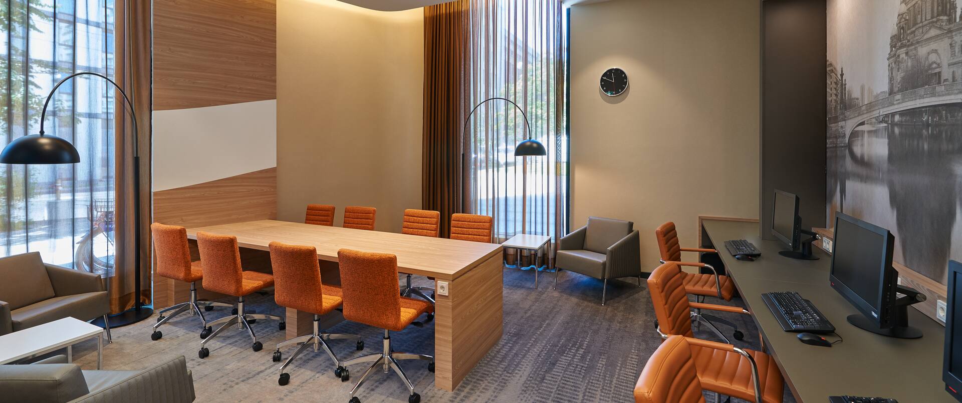 Vier Graue Sessel, zwei Stehlampen, großer Tisch mit acht orangefarbenen Stühlen, Fenster mit bodenlangen Gardinen, Wandgemälde und Schreibtisch mit drei Computern im Arbeitsbereich