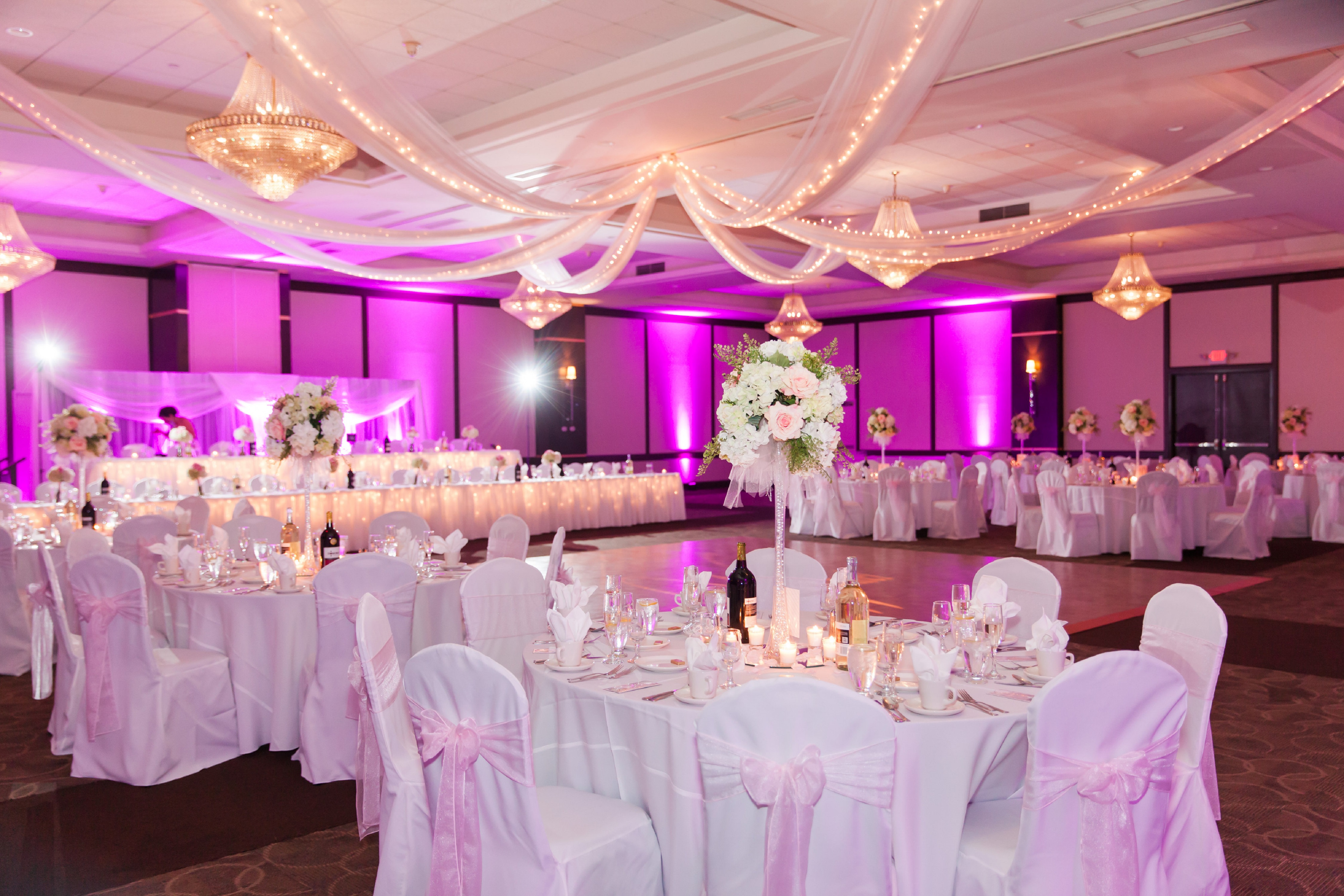 Ballroom With Pink Lighting, Place Settings, Flowers, Candles and Wine Bottles on Round Tables With White Tablecloths, and Dance Floor Set Up for Wedding Reception