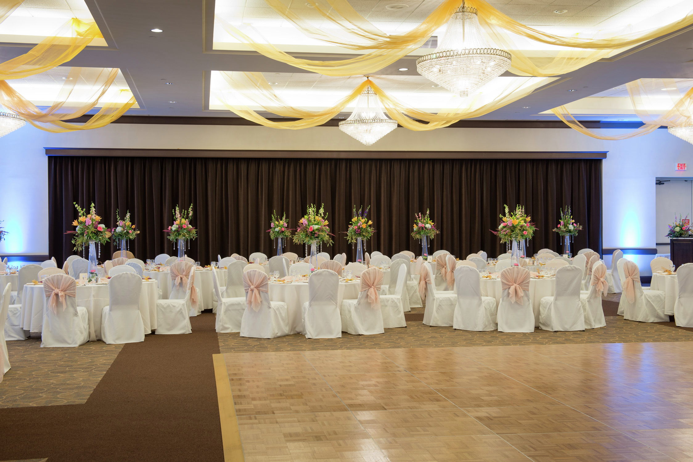 Ballroom With White Chairs, Flowers, and White Linens on Banquet Tables by Stage and Dance Floor