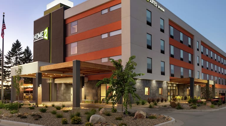 Modern Home2 Suites hotel exterior featuring porte cochere, glowing lobby, patio, and beautiful dusk sky.