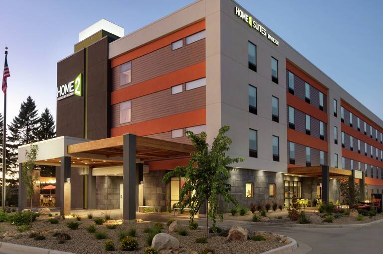 Modern Home2 Suites hotel exterior featuring porte cochere, glowing lobby, patio, and beautiful dusk sky.