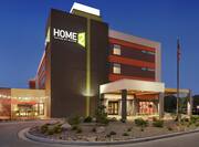 Modern Home2 Suites hotel exterior featuring porte cochere, american flag, glowing lobby, and patio with beautiful string lights.