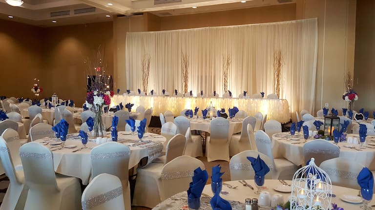 Wedding Set Up in Event Room With Place Settings, Flowers, Purple Decor, and White Linens on Round Tables, and White Chair Covers With Rhinestone Accents