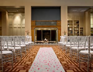 Wedding Ceremony Setup With Rose Petals on White Aisle Leading to Altar at Fireplace Surrounded by White Chairs