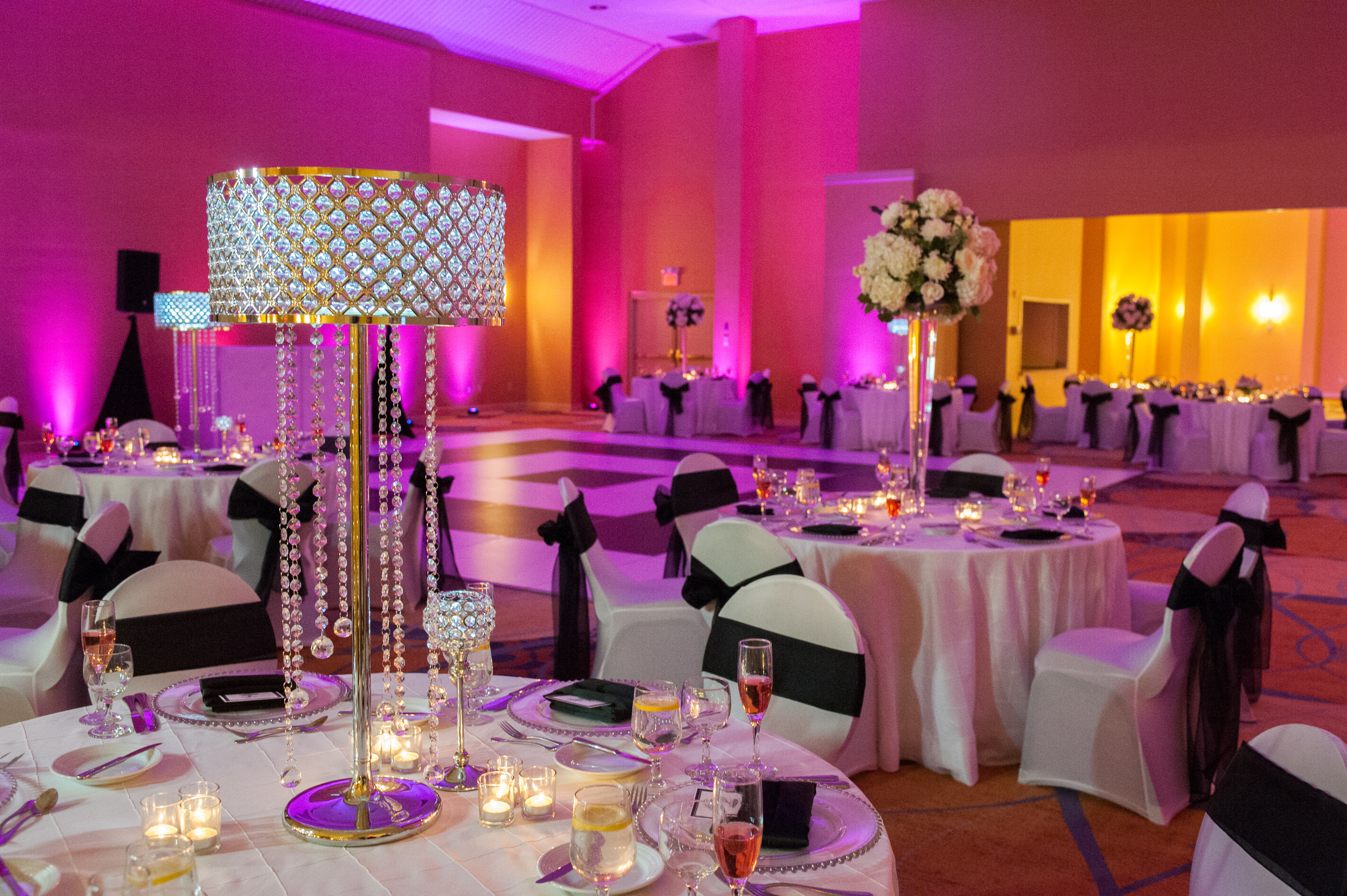  Wedding Reception With Dramatic Pink Lighting, Dance Floor, Chairs With White Covers and Black Sashes, Round Tables With Place Settings, Jeweled Lamps, White Flowers, and Candles on White Linens