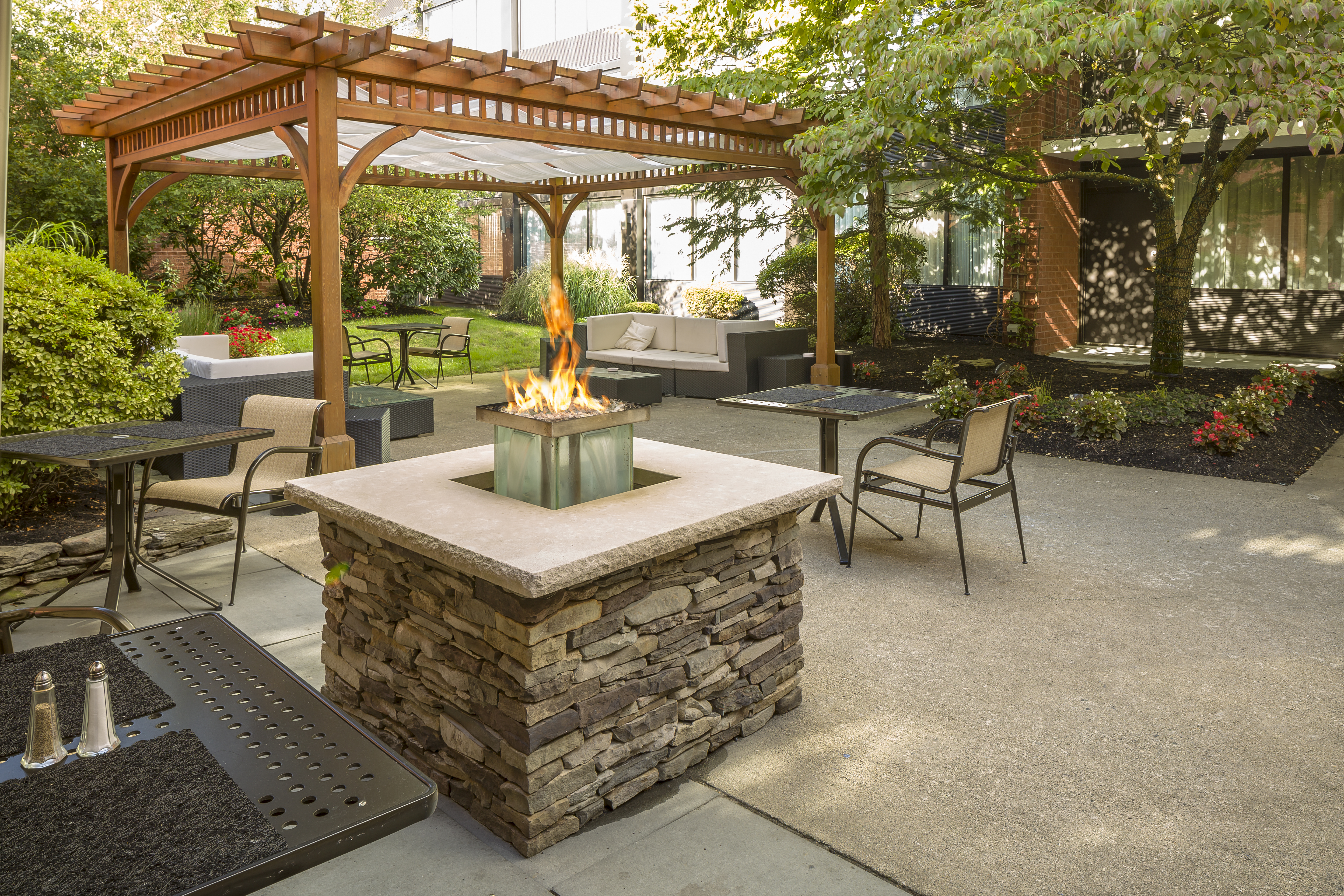  Tables and Chairs Around Fire Pit and Pavilion on Hotel Exterior Patio Surrounded by Trees
