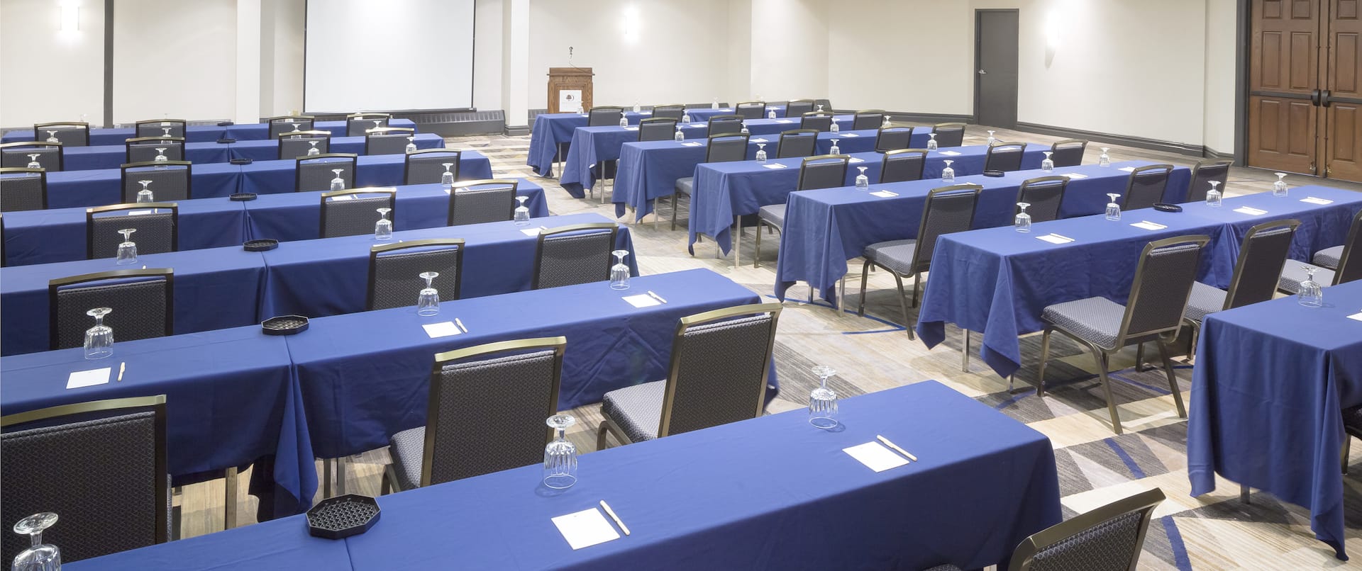 Classroom Setup in Meeting Space With Tables Covered in Blue Linens and Chairs Facing Presentation Screen and Podium