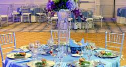 Grand  Ballroom Set Up for Wedding Reception With Dramatic Purple Lighting, Plates of Food, and Purple Flowers on Round Tables With Blue Linens and Chairs by Dance Floor