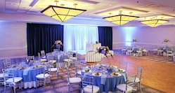 Grand Ballroom Set Up for Wedding Reception With Dramatic Purple Lighting, Head Table on a Stage, Cake Table By Dance Floor, Plates of Food, and Purple Flowers on Round Tables With Blue Linens and Chairs