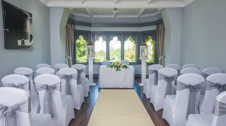 White Chairs With Grey Bows Arranged Theater Style, White Carpet Down the Center Aisle Leading to Table With White Flowers, Two Chairs, Candles and Large Window in Turret Room Set Up for Wedding Ceremony