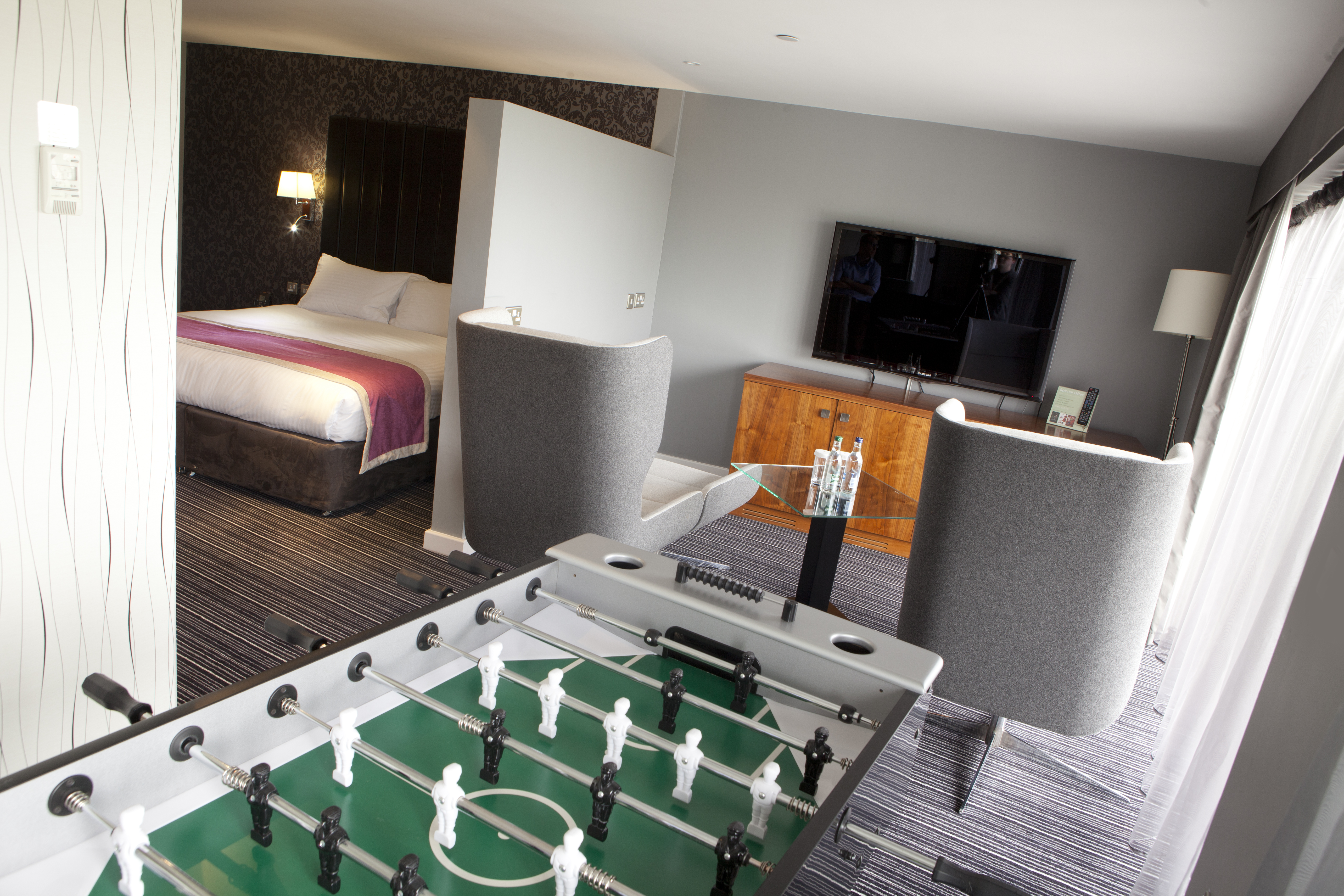  Panoramic Suite With Illuminated Lamp by Bed Behind Partition, Living Area With TV, Two Grey Armchairs, and Sheer Drapes by Balcony With a View, and Foosball Table
