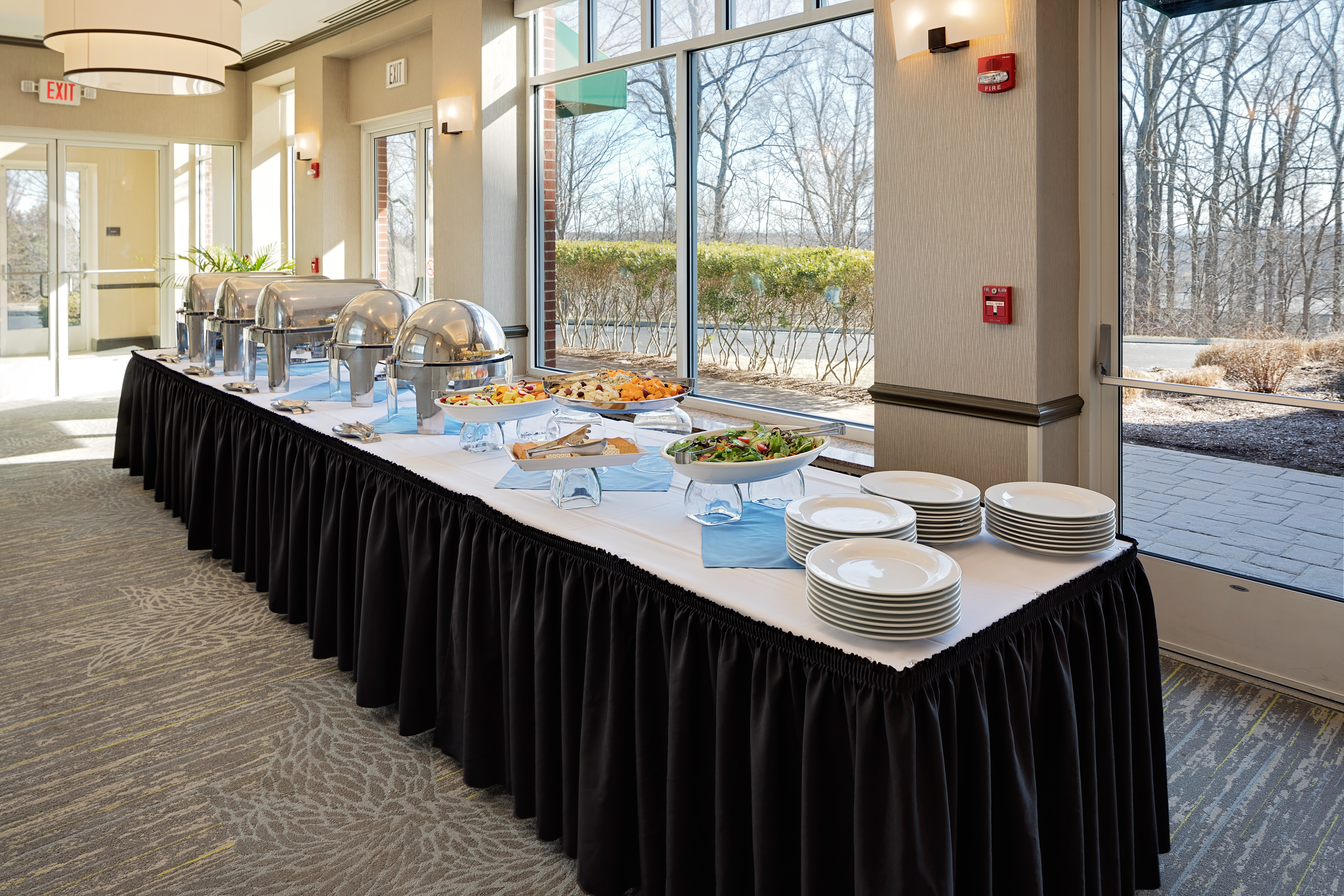 Buffet table with hot food, plates and salad as the spread. the table sits in front of large windows. 