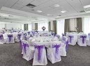 Meeting Room Set Up for Wedding Reception With Large Windows, White Chairs With Purple Bows, and Round Tables With Place Settings and Flowers on White Linens