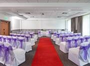 White Chairs With Purple Bows Arranged Theater Style, Red Carpet Down the Center Aisle Leading to Two Chairs in Front of Table With White Flowers, and Large Window in Meeting Room Set Up for Wedding Ceremony