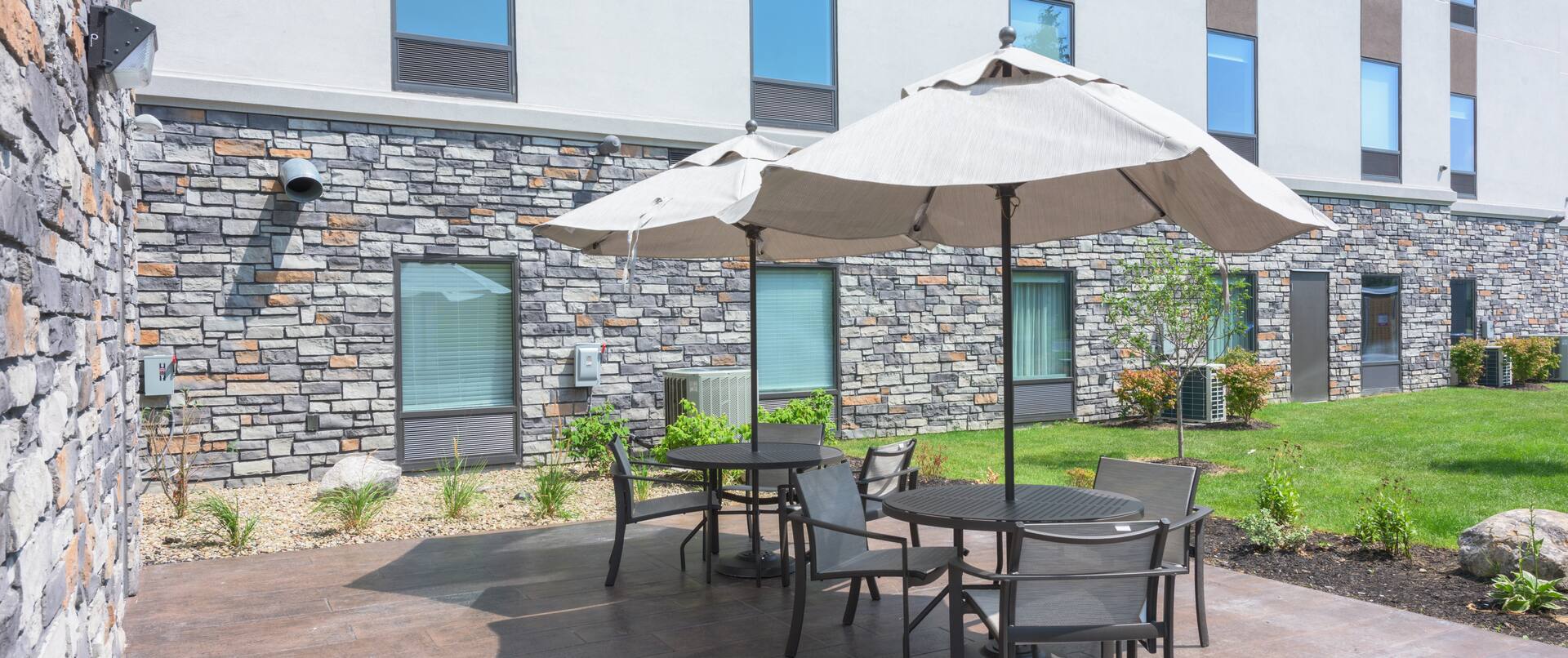 Two Patio Tables with Three Chairs and One Umbrella at Each  with view of Building Exterior and Landscaping