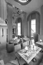 Dining lounge with small marble-top tables, upholstered chairs, arched windows, and floor-to-ceiling drapes