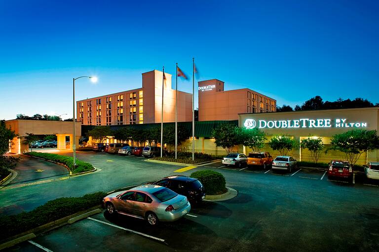 Illuminated Hotel Exterior, Signage, Porte Cochere, Flagpoles, Landscaping, and Guest Cars on Parking Lot at Night