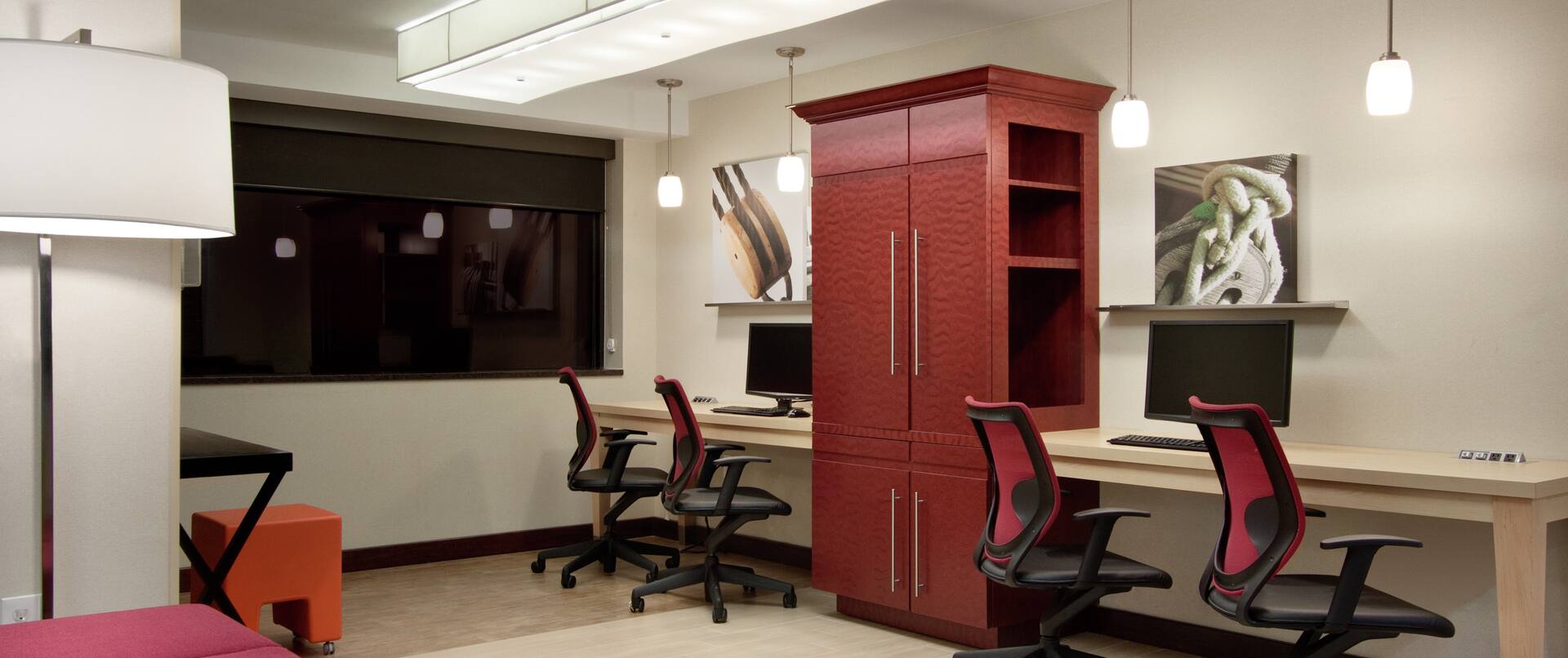 Wall Art, Red Storage Cabinet Between Two Computers on Long Desk, Four Ergonomic Chairs, and Lounge Seating With Illuminated Floor Lamp in Business Center