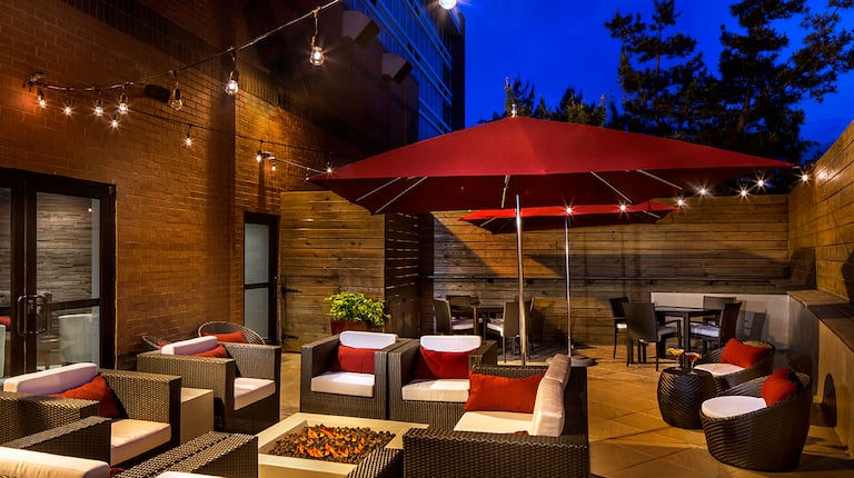 Tables With Red Umbrellas, Chairs, and Fire Pit Under Illuminated String Lights on Hotel Exterior Patio at Night