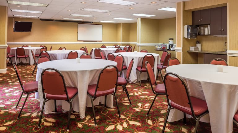 Red Chairs at Banquet Tables With White Linens, TV, Presentation Screen, and Refreshment Area in Meeting Room