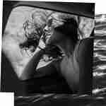 A woman in dark sunglasses sticks her head out a car window and her hair blows wildly around her. 