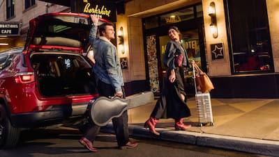 Ein Mann schließt den Kofferraum eines roten SUV, während eine Frau mit einem silbernen Koffer und einer Ledertasche auf den Bordstein tritt.