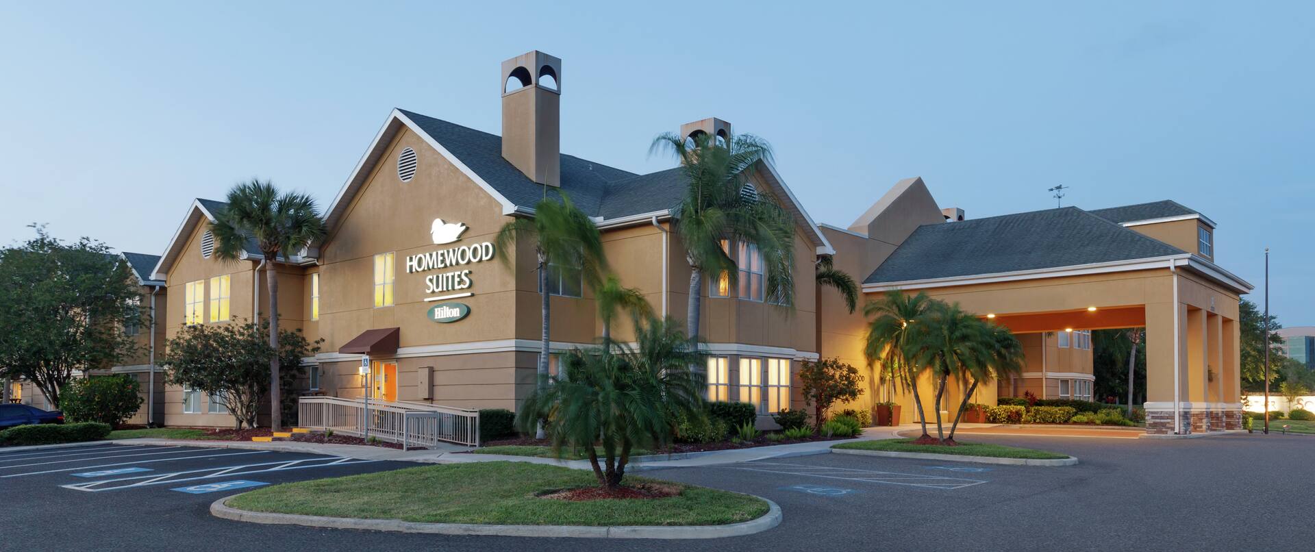 Welcoming Homewood Suites hotel exterior featuring glowing guestroom lights, beautiful palm trees, and dusk sky.
