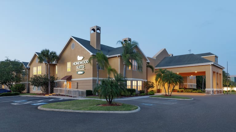 Welcoming Homewood Suites hotel exterior featuring glowing guestroom lights, beautiful palm trees, and dusk sky.