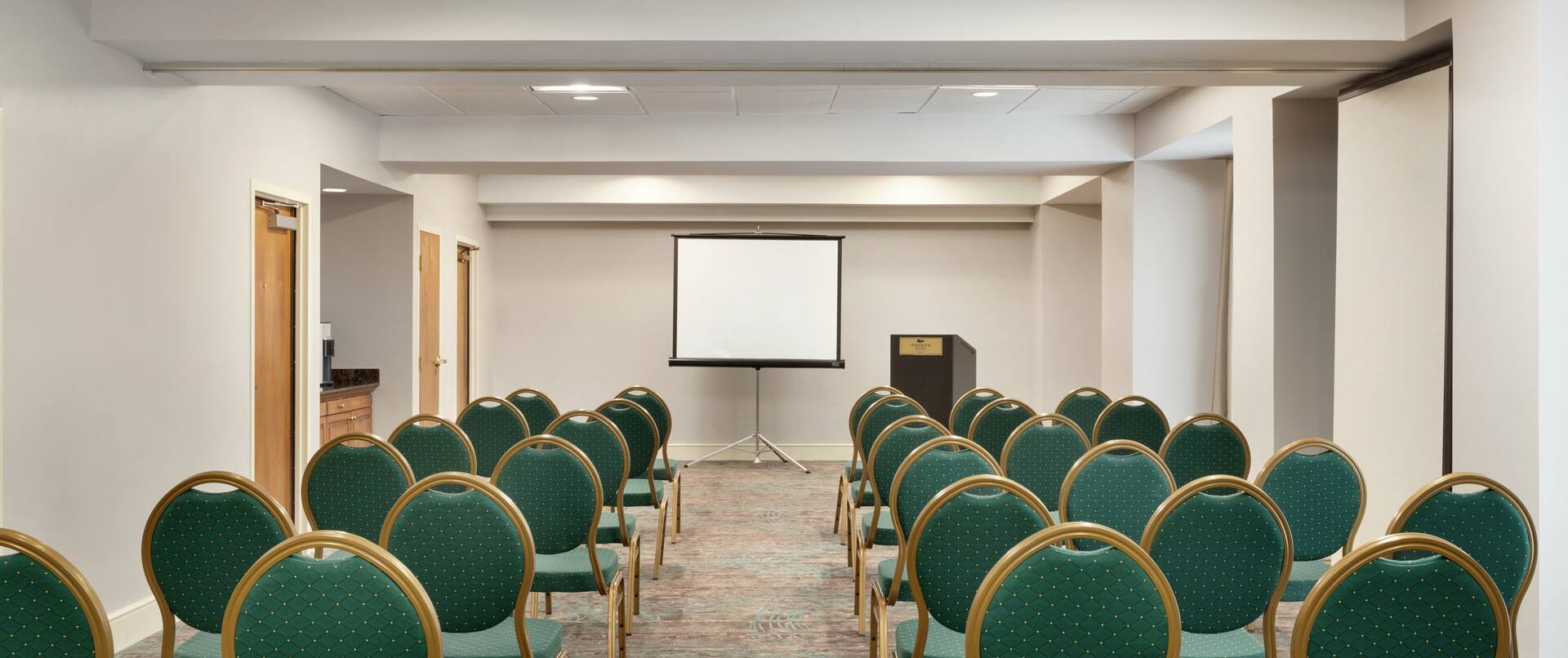 Spacious meeting room fully set in theater style with ample seating, projector screen, and podium at front of room.