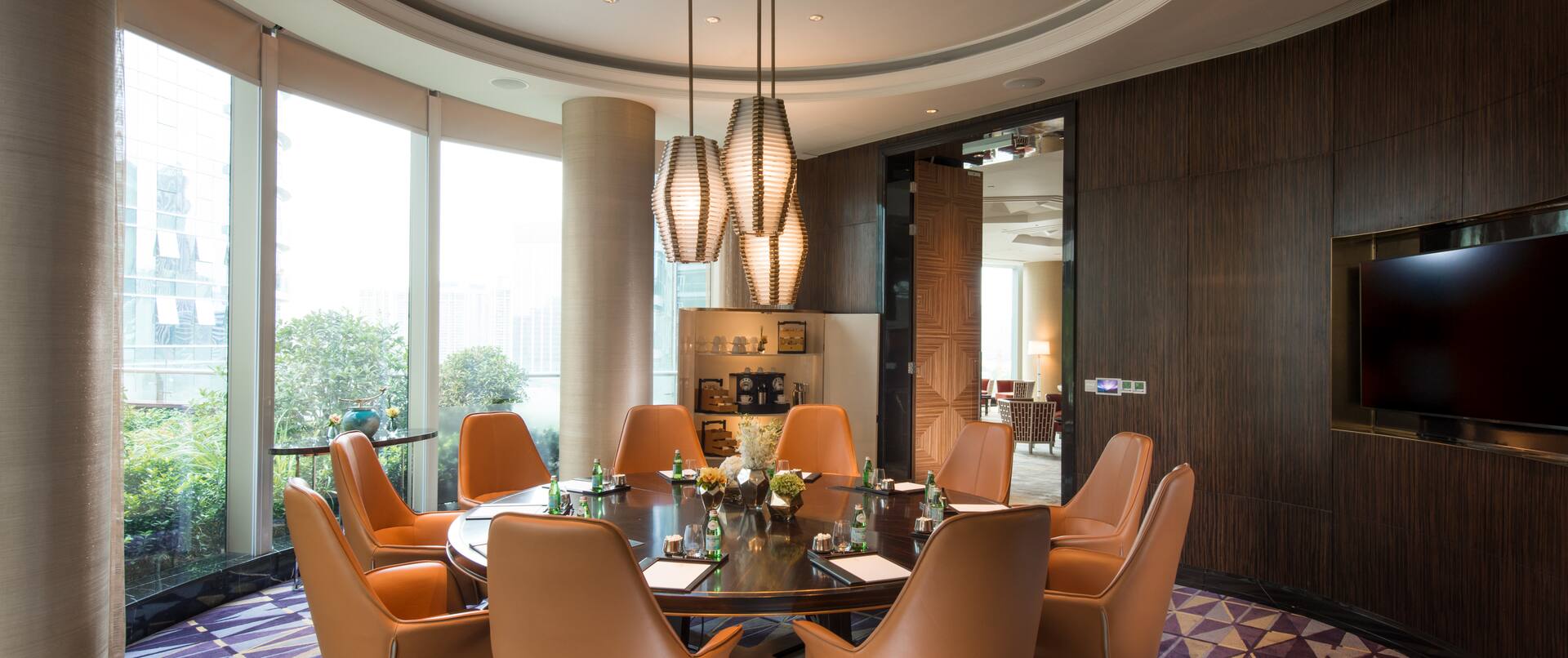 Boardroom with large round table, chairs, TV, and floor-to-ceiling windows with outdoor view