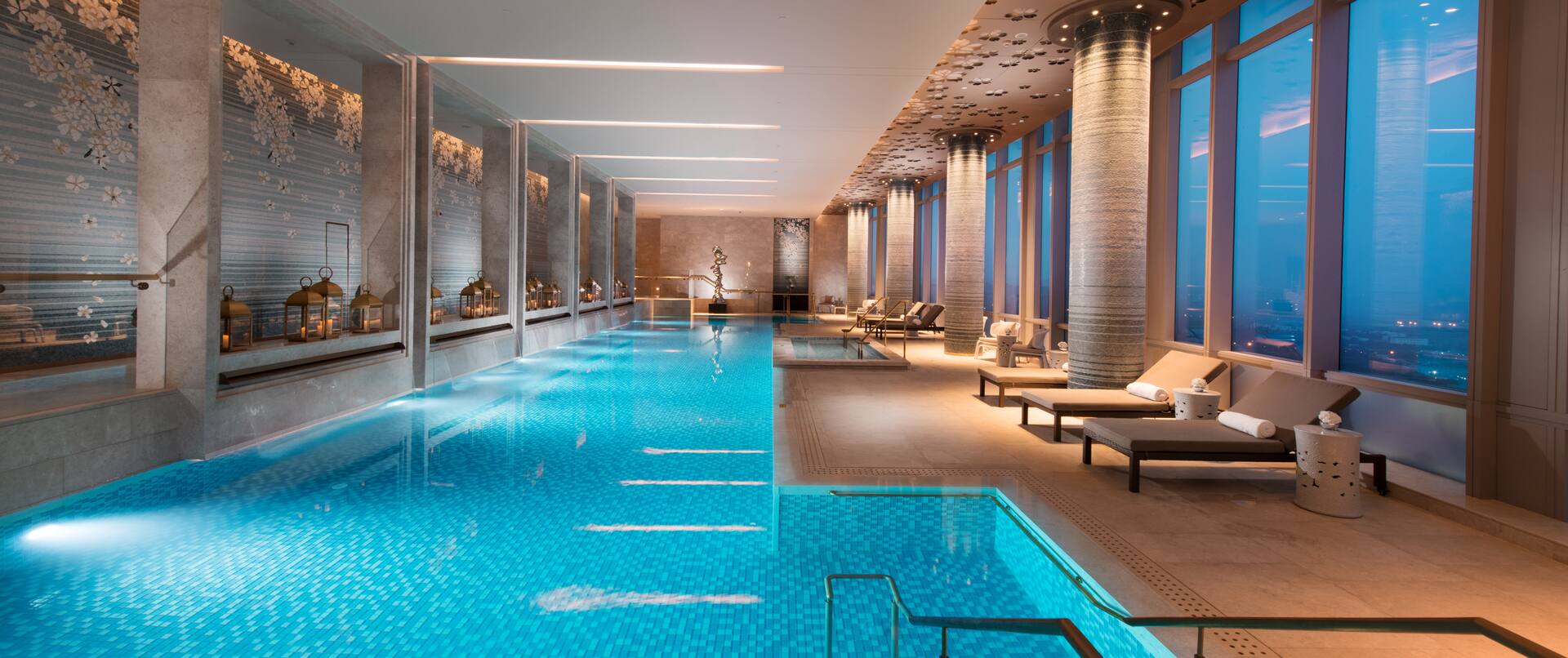 Lavish indoor pool with lounge chairs and floor-to-ceiling windows with outdoor view, lit up at night