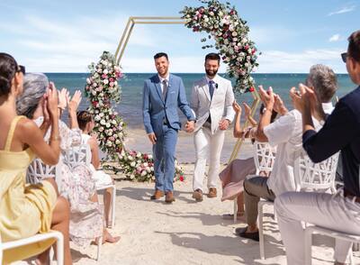 Two men in suits walk down the aisle on the beach while guests clap