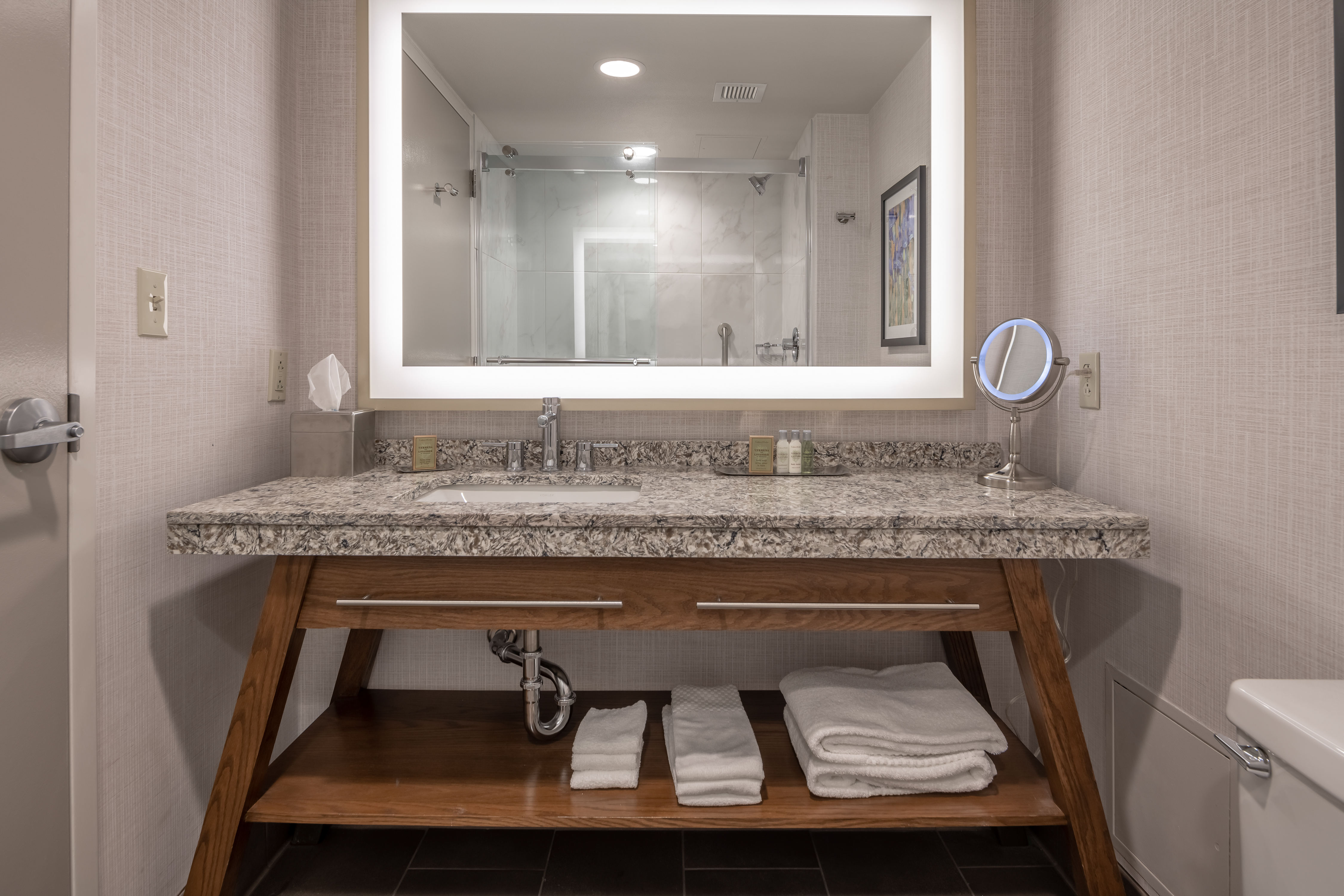 Shower With Glass Door Reflected in Brightly Lit Vanity Mirror Above Sink With Toiletries, Amenities, and Fresh Towels
