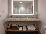 Shower With Glass Door Reflected in Brightly Lit Vanity Mirror Above Sink With Toiletries, Amenities, and Fresh Towels