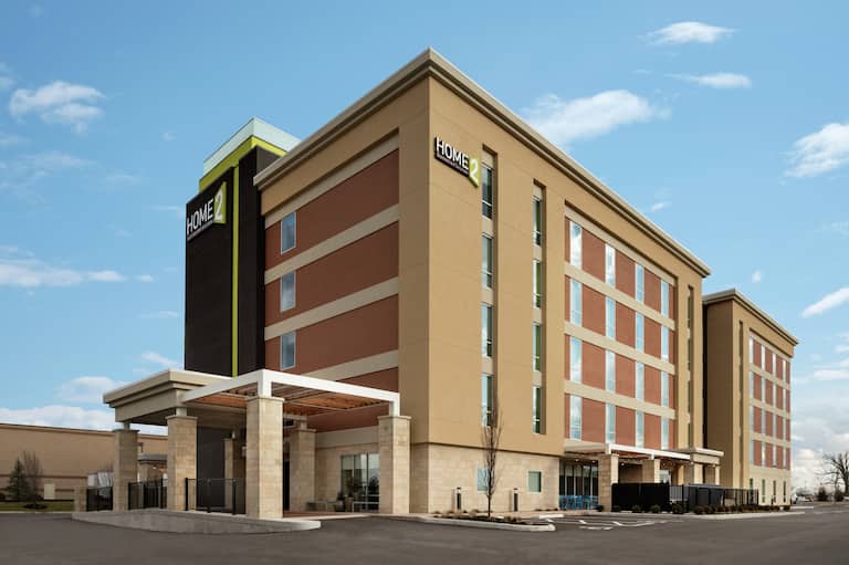 Modern Home2 Suites hotel exterior featuring porte cochere and bright blue sky.